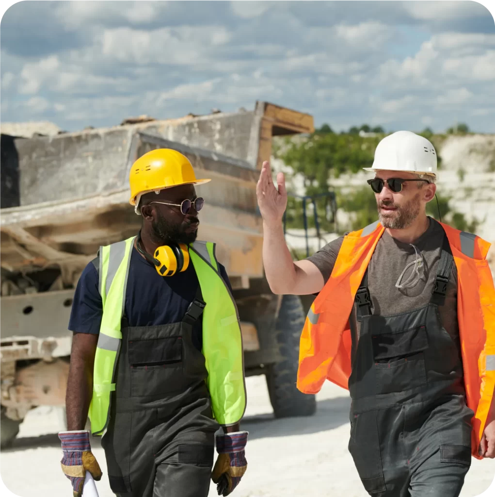 Two construction workers, wearing safety gear including helmets, vests, and gloves, walk and discuss something at a construction site with a dump truck in the background. They are reviewing aspects of their contractual liability insurance to ensure compliance and safety standards.