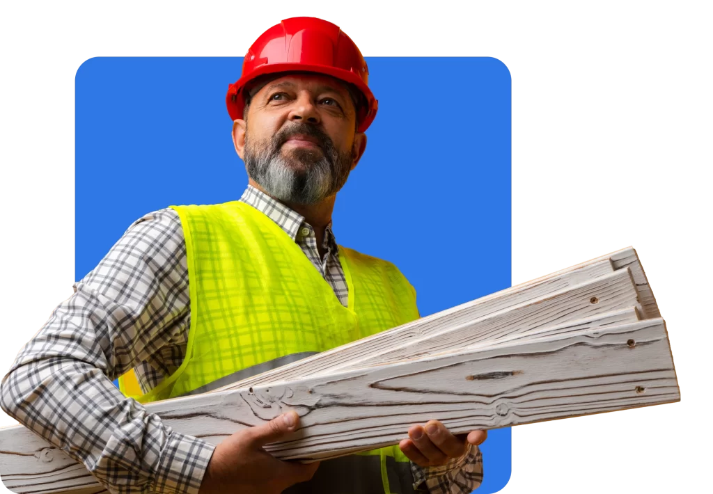 A man wearing a red hard hat and yellow safety vest holds wooden planks, highlighting the importance of builders insurance. The background is a blue rectangle.