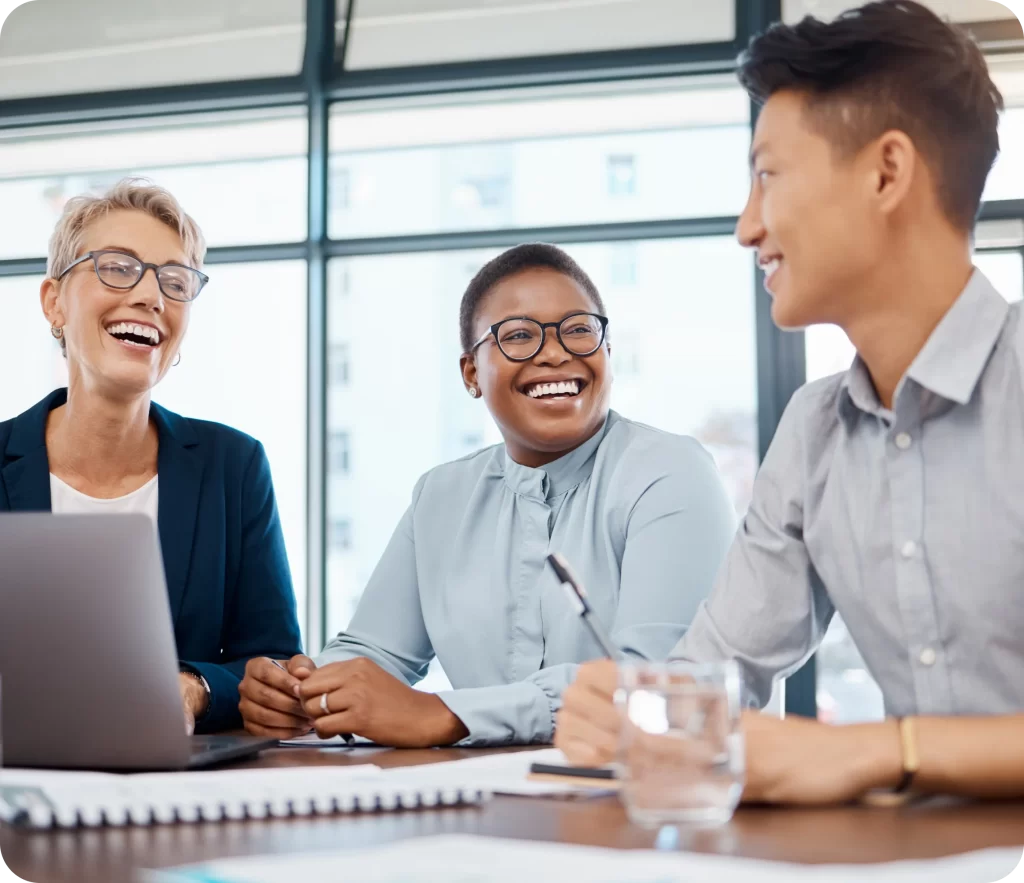 Three professionals in smart casual attire are having a discussion around a table in a bright office setting, smiling and engaged. A laptop, notebook, and glass of water are on the table.