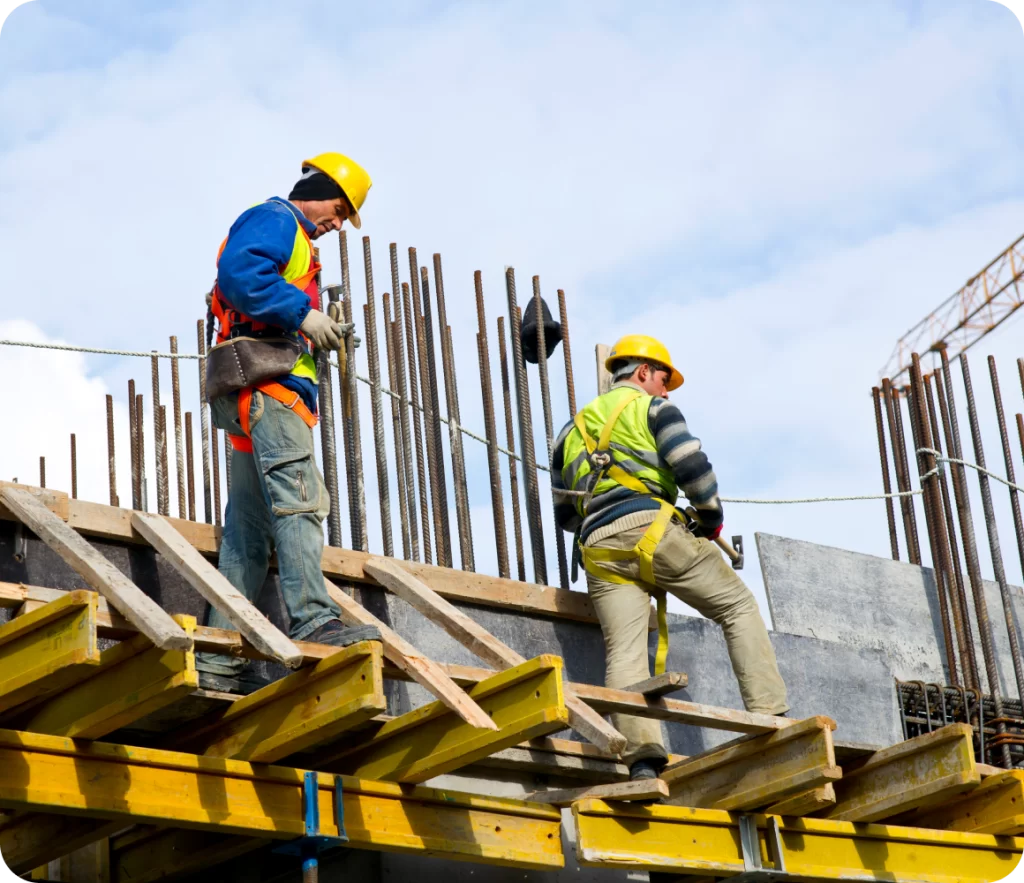 Two construction workers wearing safety gear and harnesses work on a scaffolding structure with vertical rebar rods, under a partly cloudy sky.