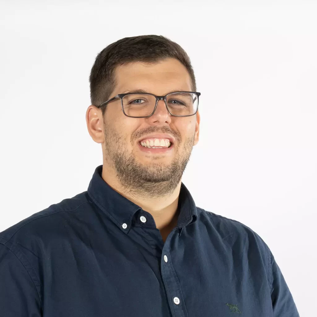 A person with glasses, short hair, and a beard wearing a dark button-up shirt, smiling against a plain white background.