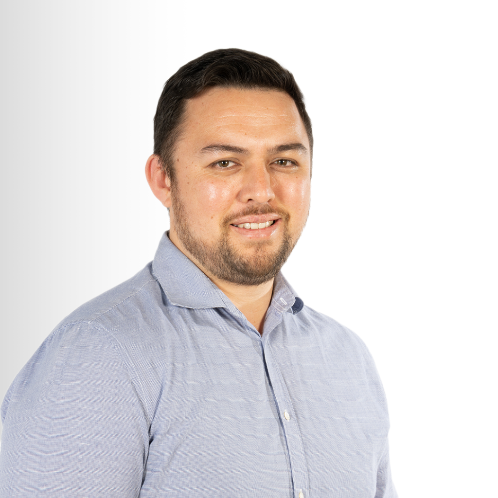 A person with short dark hair and a beard, wearing a light blue button-up shirt, is smiling and facing the camera against a white gradient background.